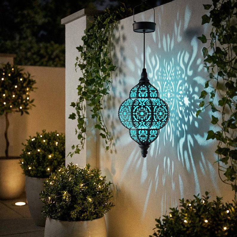 May include: A black and turquoise solar-powered lantern with an intricate, openwork design hangs from a hook. The lantern casts a decorative light pattern on the wall. Green ivy and small potted plants with fairy lights surround the lantern.