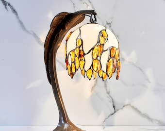 Puede incluir: Una lámpara de mesa de color bronce con una pantalla de vidrieras. La pantalla es blanca con piezas de vidrio ámbar y amarillo. La lámpara tiene un tallo curvo y una base decorativa. La lámpara está diseñada para parecerse a un pájaro.