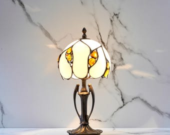 Può includere: Lampada da tavolo decorativa con paralume in vetro colorato bianco e ambra. La lampada ha una base e uno stelo color bronzo con un design ornato. Il paralume è illuminato, creando una luce calda e invitante.
