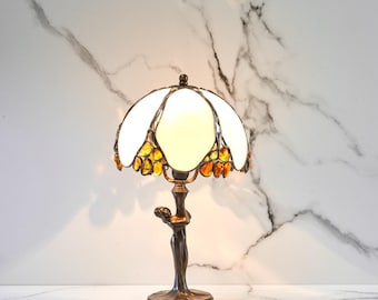 Può includere: Una lampada da tavolo decorativa con paralume in vetro colorato bianco e ambra. La lampada ha una base color bronzo con una figura che sostiene il paralume. Il paralume ha un motivo floreale ed è illuminato dall'interno.