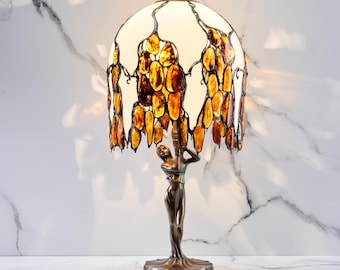 Puede incluir: Lámpara de mesa de bronce con una figura femenina que sostiene una pantalla abovedada. La pantalla es blanca con piezas de vidrio irregulares de color ámbar. La lámpara tiene una base redonda y un remate decorativo.