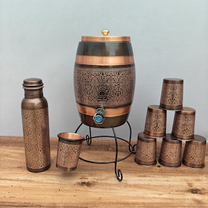 May include: A copper-toned water dispenser set with an ornate floral pattern. The set includes a barrel-shaped dispenser with a spigot, a bottle, and several small cups. The dispenser sits on a black metal stand.