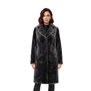 May include: A long, dark gray faux fur coat with a wide collar and a button closure. The coat is knee-length and has a soft, textured appearance. The model is wearing black pants.