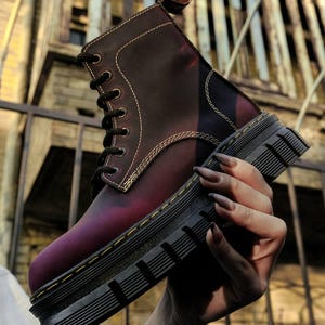 Könnte beinhalten: Ein burgunderfarbener und violetter Lederstiefel mit schwarzen Schnürsenkeln und einer dicken schwarzen Sohle. Der Stiefel hat kontrastierende weiße Nähte und eine Zuglasche oben. Der Stiefel wird vor einem verschwommenen Hintergrund gehalten.