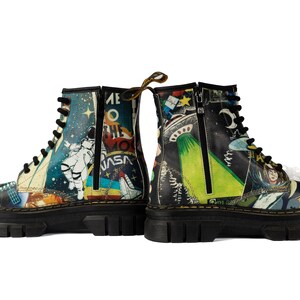 Könnte beinhalten: Paar schwarze Kampfstiefel mit farbenfrohen Weltraum-Grafiken. Ein Stiefel zeigt einen Astronauten und das NASA-Logo, der andere ein UFO und den Mond. Beide Stiefel haben schwarze Schnürsenkel, Reißverschlüsse und dicke Sohlen.