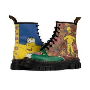 Könnte beinhalten: Paar schwarze Kampfstiefel mit den Simpsons-Charakteren. Ein Stiefel hat ein blaues und gelbes Design mit Bart Simpson, der andere ein farbenfrohes Design mit Homer Simpson und Donuts. Die Stiefel haben schwarze Schnürsenkel und Sohlen.