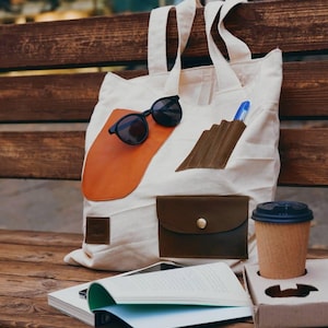 Könnte beinhalten: Cremefarbene Tragetasche mit braunen Lederdetails, Sonnenbrille und Stift. Ein Buch, ein Smartphone und eine Kaffeetasse in einem Pappbecherhalter liegen auf einer Holzbank. Die Tasche hat einen großen orangefarbenen Lederaufnäher.