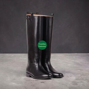 Pode incluir: Um par de botas de montaria altas em couro preto com acabamento liso e polido, salto baixo e um detalhe de tira com fivela no topo do cano. O texto Frontline Footwear está impresso em branco num emblema circular de cor verde.
