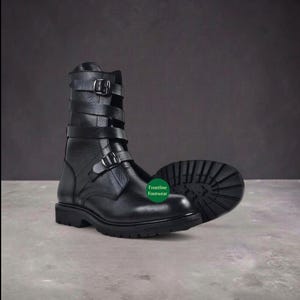 Könnte beinhalten: Ein wadenhoher Stiefel aus schwarzem Leder mit drei horizontalen Riemen mit silberfarbenen Metallschnallen am Schaft und einem vierten Riemen über dem Spann. Der Stiefel hat eine dicke, profilierte Gummisohle und ein grünes Logo mit der Aufschrift Frontline Footwear.