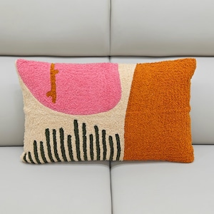 Puede incluir: Cojín decorativo rectangular con un diseño abstracto texturizado. El cojín presenta un semicírculo rosa, un rectángulo naranja y líneas verticales negras sobre un fondo crema. El cojín está colocado sobre una superficie gris claro.