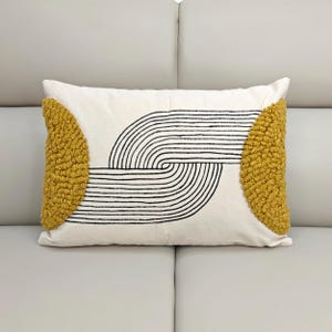 Puede incluir: Cojín decorativo rectangular con fondo crema y diseño lineal negro curvo. Presenta detalles texturizados amarillo mostaza en cada extremo. El cojín está sobre una superficie gris claro.