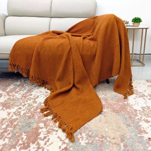 May include: An orange-brown waffle-knit throw blanket with fringed edges draped over a chair. The blanket is made of a textured material and is a warm, inviting color. The blanket is on a chair in a living room setting.