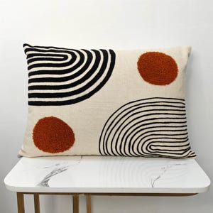 May include: Rectangular decorative pillow with a cream-colored background. The pillow features black curved lines and two rust-colored circles. The pillow is resting on a white marble table with gold legs. The pillow is a decorative home accent.
