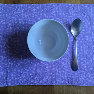 Peut inclure: Un set de table rectangulaire lavande avec un motif floral blanc répétitif, surmonté d'un bol en céramique blanche et d'une cuillère en acier inoxydable. Une petite étiquette en tissu blanc est fixée au coin inférieur gauche du set.