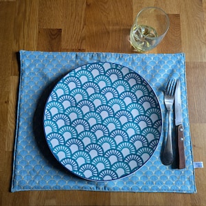Peut inclure: Un set de table bleu avec un motif doré, une assiette avec un motif en éventail turquoise et blanc, des couverts et un verre de liquide sur une table en bois. L'assiette est centrée sur le set de table.