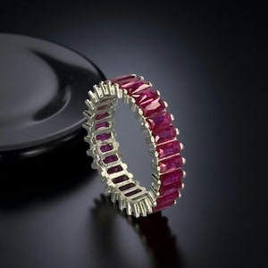 Puede incluir: Un anillo de plata con una banda continua de piedras preciosas rectangulares de color rojo intenso. El anillo se muestra sobre un fondo oscuro, resaltando el brillo de las piedras. Una joya.