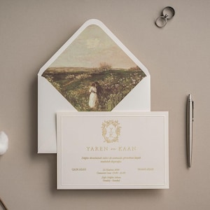 Puede incluir: Una tarjeta de invitación de boda color crema con texto dorado YAREN ve KAAN y un sobre a juego con una pintura de paisaje de una pareja en un campo. Incluye dos anillos plateados, un bolígrafo plateado y una rama de flor de algodón.
