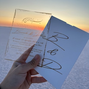 May include: A clear acrylic wedding invitation with gold calligraphy, held against a sunset. The invitation includes names, date, time, and location details. A white envelope with gold calligraphy is also visible.