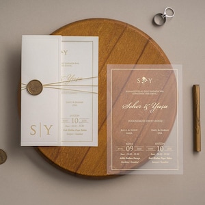 Könnte beinhalten: Ein Hochzeitseinladungsset bestehend aus einer weißen Karte mit goldener Schrift, einem Wachssiegel und goldener Schnur, sowie einer transparenten Acrylkarte mit dem weißen Text Seher & Yusa, zwei Metallringen und zwei bronzefarbenen Münzen.