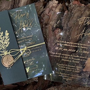 Puede incluir: Elegantes invitaciones de boda con letras y detalles dorados. Las tarjetas verde oscuro presentan diseños florales y un sello de cera. La invitación incluye los nombres Flavie & Abdel y los detalles del evento.