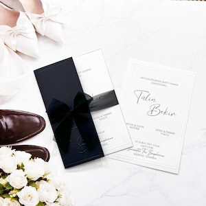 May include: A wedding invitation set featuring a dark navy blue card with a black ribbon bow and a white card with black text reading Tülin Bekin, 25 Şubat 2027 Pazar, and Swissotel The Bosphorus, accompanied by white satin shoes and white roses.