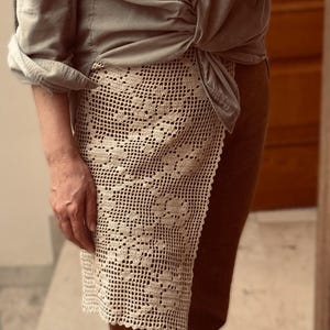 May include: A cream-colored, openwork crochet skirt with a floral pattern. The skirt is draped over brown pants. A gray button-down shirt is tied at the waist. The image is taken from the waist down.