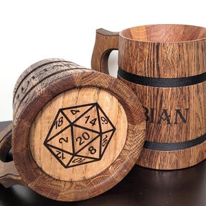 Op de afbeelding: Twee houten bierpullen. Eén pul ligt op zijn kant en toont een zwart-wit D20-dobbelsteenafbeelding. De andere staat rechtop, met de naam "BIAN" op de zijkant gegraveerd. Beide pullen hebben een donkere houtnerf afwerking.