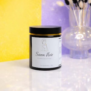 May include: A black jar with a gold rim and a black lid, labelled "Savon Noir." The label features an illustration of a woman and text in French. The jar contains 250g of product. The background is a mix of yellow, purple, and white.