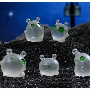 Peut inclure: Cinq petites figurines translucides en blanc dépoli avec des corps arrondis et des oreilles. Chaque figurine présente des yeux circulaires vert vif et des expressions faciales simples, notamment des clins d'œil, des sourires et des regards neutres.
