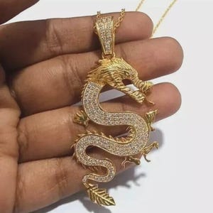 Könnte beinhalten: Eine goldfarbene Drachen-Anhänger-Halskette. Der Drache ist mit kleinen, klaren Edelsteinen besetzt und hat ein detailliertes, strukturiertes Design. Der Anhänger hängt an einer zarten Goldkette.