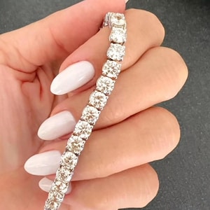 Puede incluir: Una pulsera de tenis de diamantes. La pulsera presenta una serie de diamantes de talla redonda engarzados en una fila. Los diamantes son claros y brillantes. La pulsera se sostiene sobre un fondo neutro.