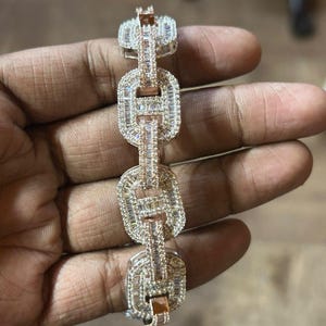 Könnte beinhalten: Ein roségoldenes und silbernes Armband mit einem Kettenglied-Design. Die Glieder sind rechteckig und mit kleinen, funkelnden Steinen besetzt. Das Armband wird in einer Hand gehalten.