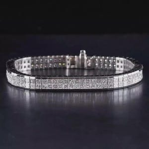May include: A silver bracelet with two rows of square-cut, clear gemstones. The bracelet has a secure clasp and is displayed against a dark background. The gemstones are set closely together, creating a continuous sparkle.