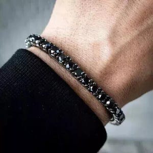 May include: A black gemstone bracelet with a row of square-cut black stones. The bracelet is made of a silver-colored metal and is worn on a wrist. The background is a blurred view of a person's arm and clothing.