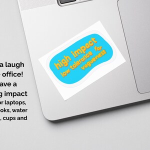May include: A sticker on a laptop with the text "high impact low tolerance for vagueness" in orange on a blue background. The sticker is white-bordered and is on a silver laptop.