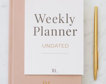 Könnte beinhalten: Ein rosafarbener Wochenplaner mit dem Text "Weekly Planner UNDATED". Der Planer hat die Initialen "BL" auf dem Einband geprägt. Ein goldener Stift und zwei goldene Büroklammern liegen neben dem Planer auf einer Marmoroberfläche.