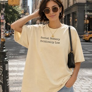 Woman wearing a cream t-shirt with "Social Battery Critically Low" text.