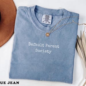 以下が含まれることがあります： 「Default Parent Society」の白文字がプリントされた、ブルーデニムのComfort Colors Tシャツ。ペンダント付きのゴールドネックレスがシャツの上にあります。茶色の帽子と乾燥した植物が背景にあります。