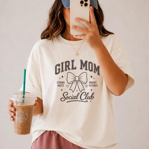 Woman in cream t-shirt with "Girl Mom Social Club" text, holding iced coffee.