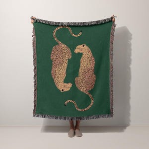 May include: A green throw blanket featuring two leopard illustrations in tan and brown. The blanket has a fringed edge in black and pink. The leopards are facing each other, with one upright and the other upside down.