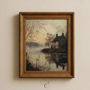 May include: A framed landscape painting depicts a serene waterside scene. A small stone house with a smoking chimney sits on the bank of a calm lake. Reeds and trees line the shore, and the sky is a soft blend of colors. The painting is in a wooden frame.