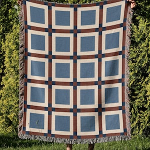 May include: A woven throw blanket featuring a repeating geometric grid pattern of blue squares framed by cream borders and dark brown intersecting lines. The rectangular textile has a fringed edge along the top and bottom, showcasing a multi-colored thread construction throughout the entire surface of the fabric.