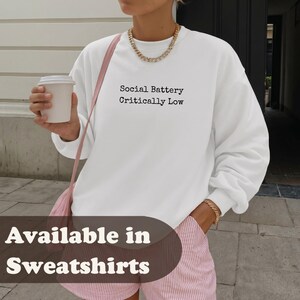 Woman wearing a white sweatshirt with "Social Battery Critically Low" text.