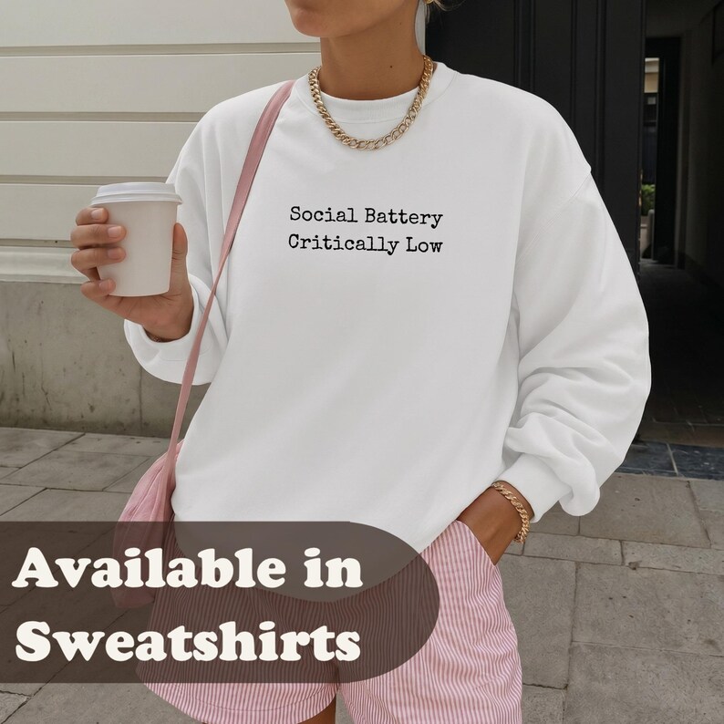 Woman wearing a white sweatshirt with "Social Battery Critically Low" text.