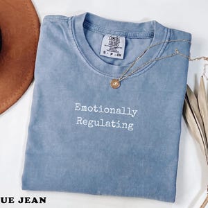 Può includere: Una maglietta blu jeans con le parole "Emotionally Regulating" in bianco. Una collana d'oro con un ciondolo è drappeggiata sulla maglietta. La maglietta è piegata e ha un'etichetta con la scritta "Comfort Colors".
