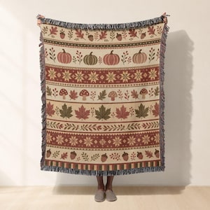 May include: A woven throw blanket featuring a repeating horizontal pattern of pumpkins, mushrooms, maple leaves, acorns, and floral sprigs in shades of orange, brown, green, and cream, finished with a decorative fringe along the top and bottom edges.