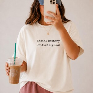 Cream t-shirt with "Social Battery Critically Low" text, held by person with iced coffee.