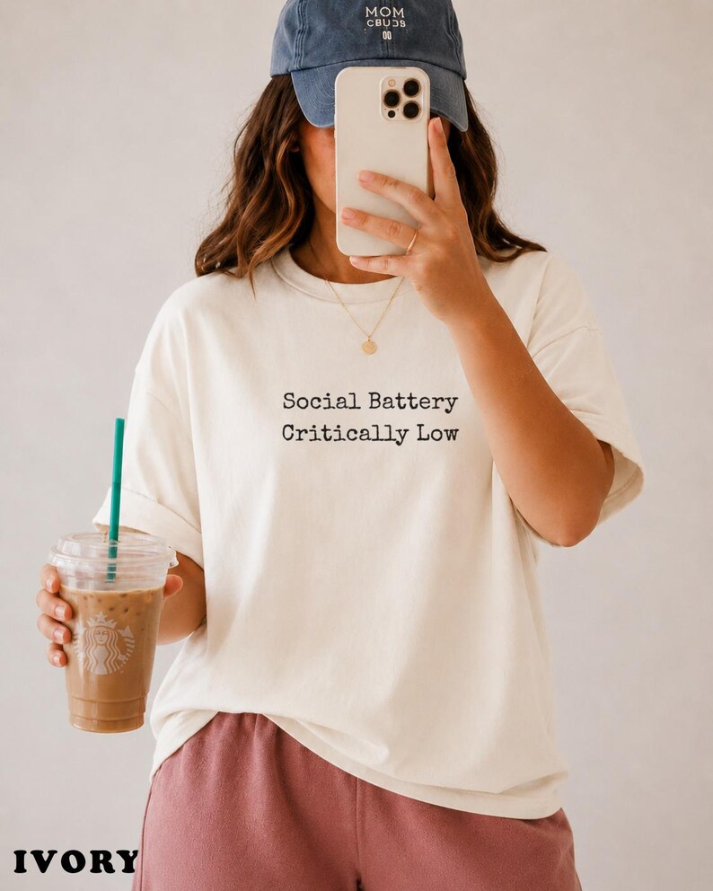 Cream t-shirt with "Social Battery Critically Low" text, held by person with iced coffee.