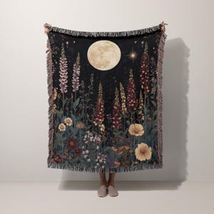 May include: A black woven throw blanket featuring a nighttime floral scene with a full moon, stars, and various flowers in shades of purple, red, and yellow. The blanket has a fringed edge.