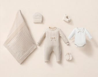 May include: A collection of cream-colored baby clothing items, including a blanket, hat, romper, bodysuit, booties, and mittens. The blanket features a cable knit design. The romper has a bow detail. The items are arranged on a light beige background.